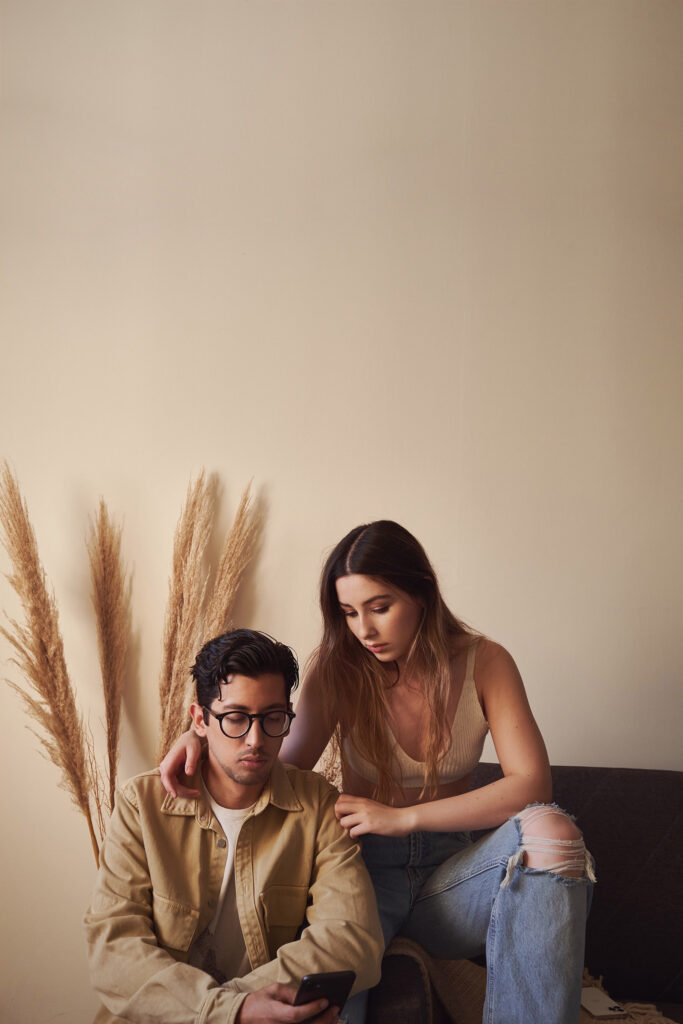 Gaby and Me a couple sit against a beige wall with tall dried grass. Gaby with long brown hair leans over Mehadi who wears glasses and holds a phone.